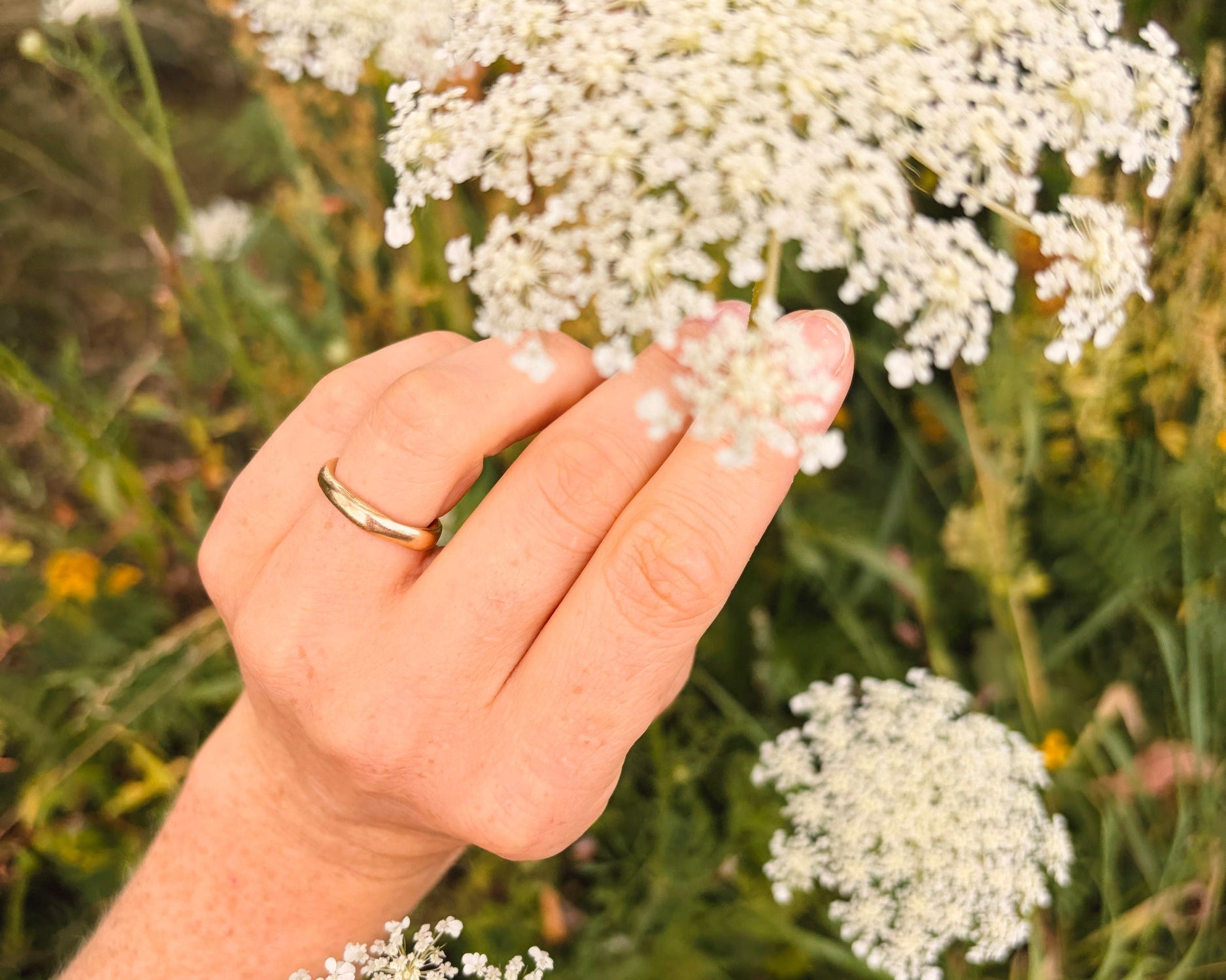 Thin yellow gold wedding band worn on a hand beside white flowers