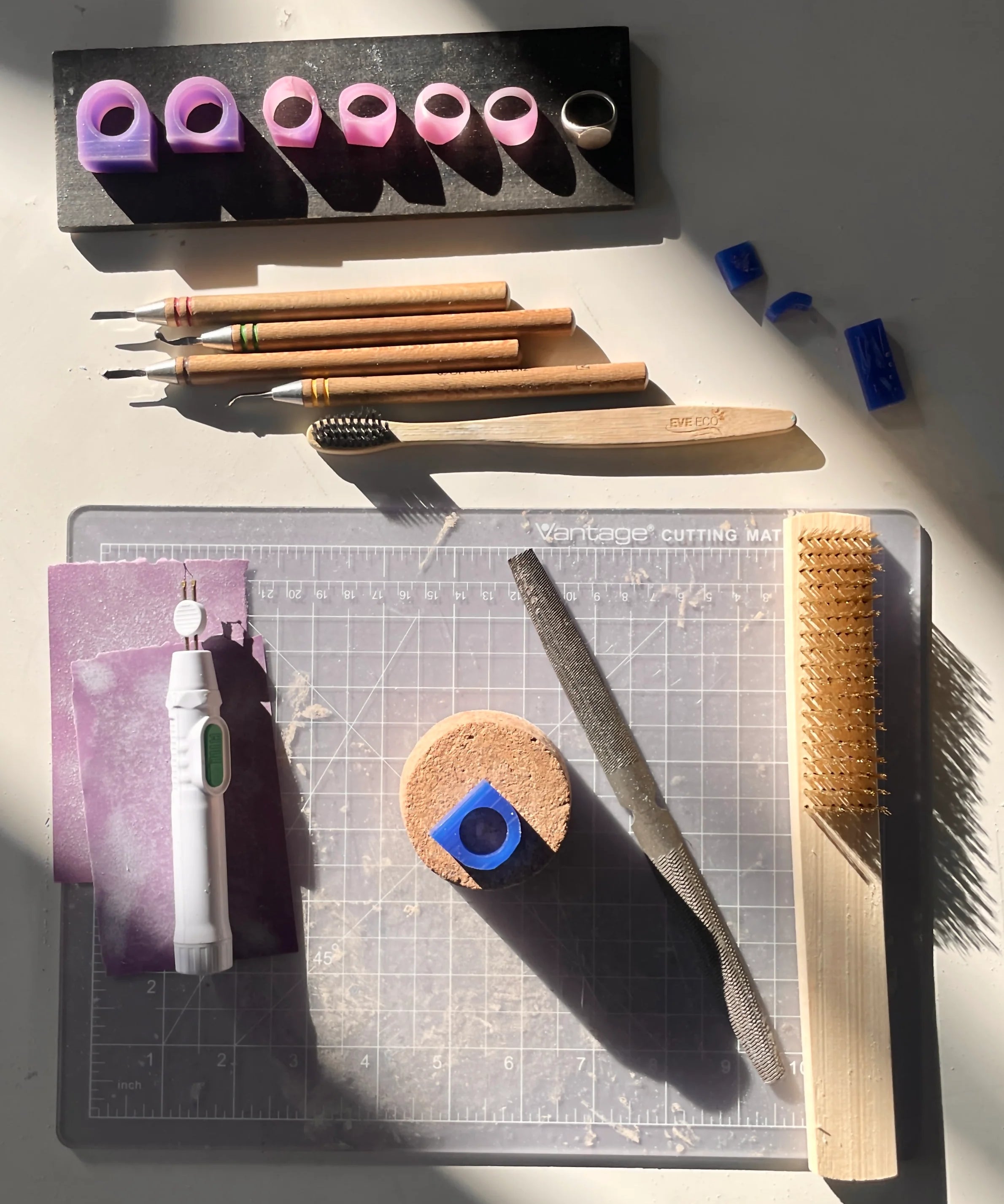 Collection of wax carving jewelry tools and materials on a cutting mat with a gray background