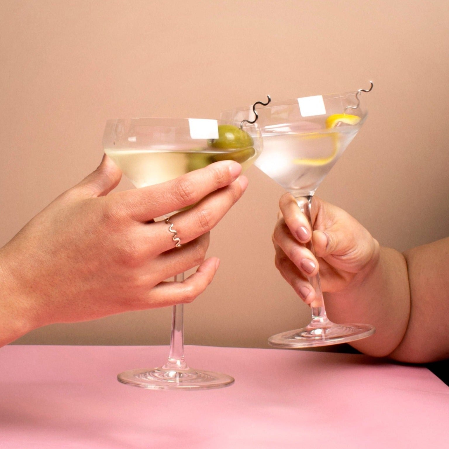 two hands holding martini glasses toasting. Both martinis have curvy sterling silver cocktail picks, one holds a lemon peel, the other three olives.  one hand wears a curvy ring.  the background is tan on top and pink on the bottom.